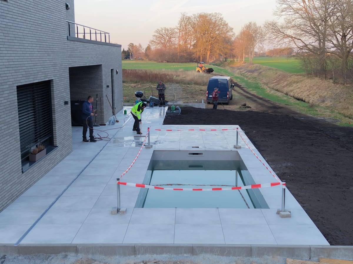 Abendaufnahme der Baustelle: verlegte Terrassenplatten um das abgesperrte Becken, zwei Arbeiter am Beckenrand, daneben offener Mutterboden