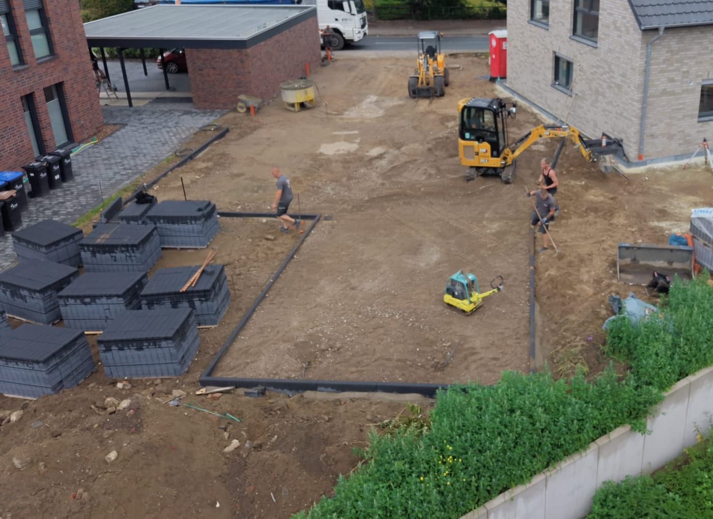 Luftaufnahme einer Hofflaeche im Bau: gestapelte Rigolenbloecke fuer die Regenwasserversickerung, Minibagger, Ruettelplatte und abgezogenes Planum